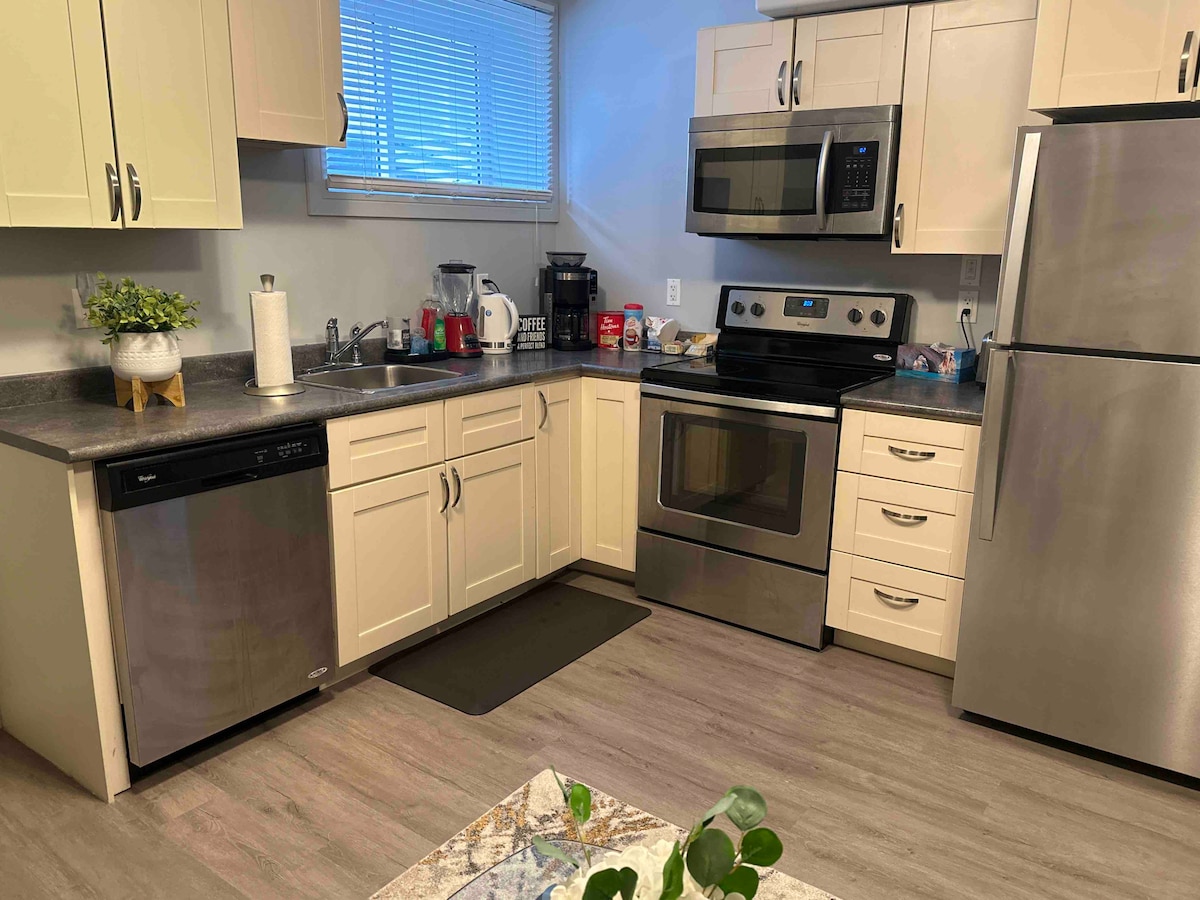 The kitchen features modern appliances, including a microwave and stainless steel refrigerator. Light-colored cabinets provide ample storage. A dishwashing machine is positioned next to the sink, and a countertop offers workspace. Natural light enters through a nearby window, creating an inviting atmosphere.
