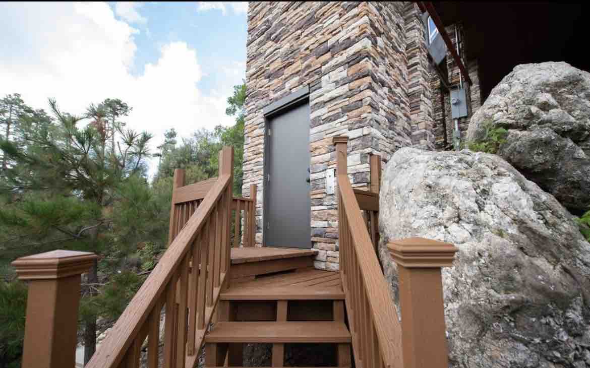 A wooden staircase leads to a solid door, framed by natural stone walls. Surrounding the entrance, large rocks and greenery create a rustic outdoor setting, with steps designed for easy access to the cabin.