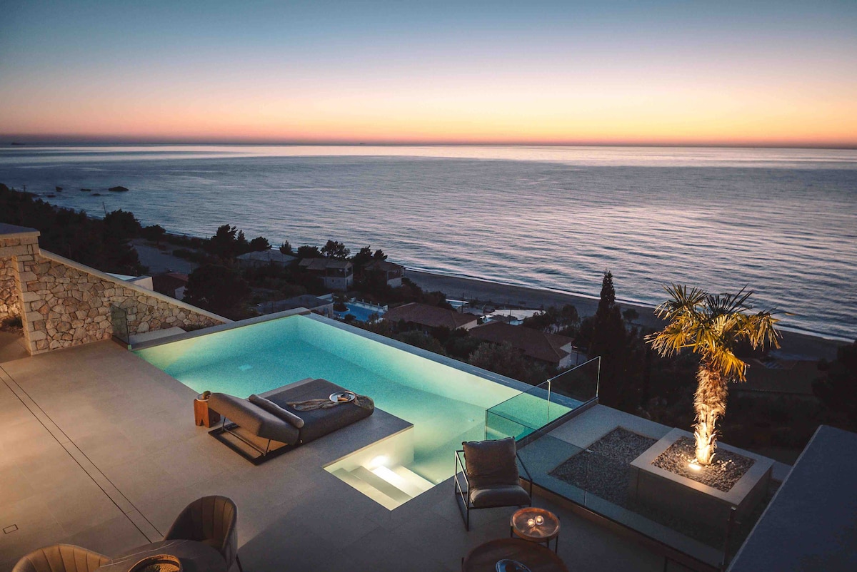 An infinity pool is positioned at the edge of a terrace, offering unobstructed views of the ocean and sunset. Lounge chairs are placed nearby, while a palm tree adds a touch of greenery. Soft lighting illuminates the area, contributing to a serene atmosphere.