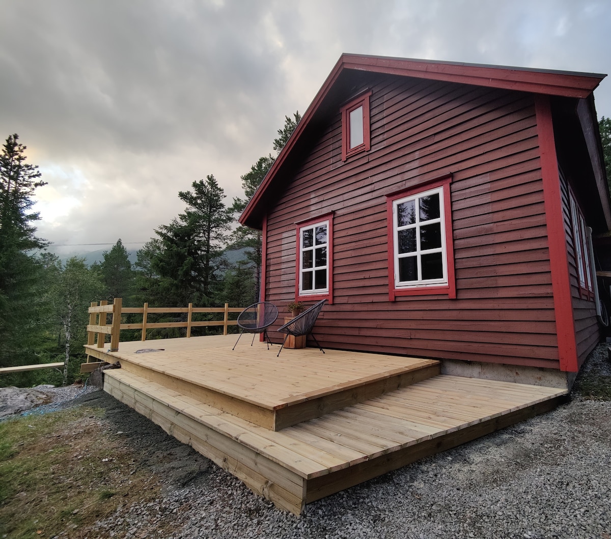 The cabin exterior is characterized by traditional red siding and white-framed windows. A spacious wooden deck is featured in front, with two black chairs positioned for outdoor relaxation. Surrounding trees enhance the natural setting, creating a serene atmosphere.