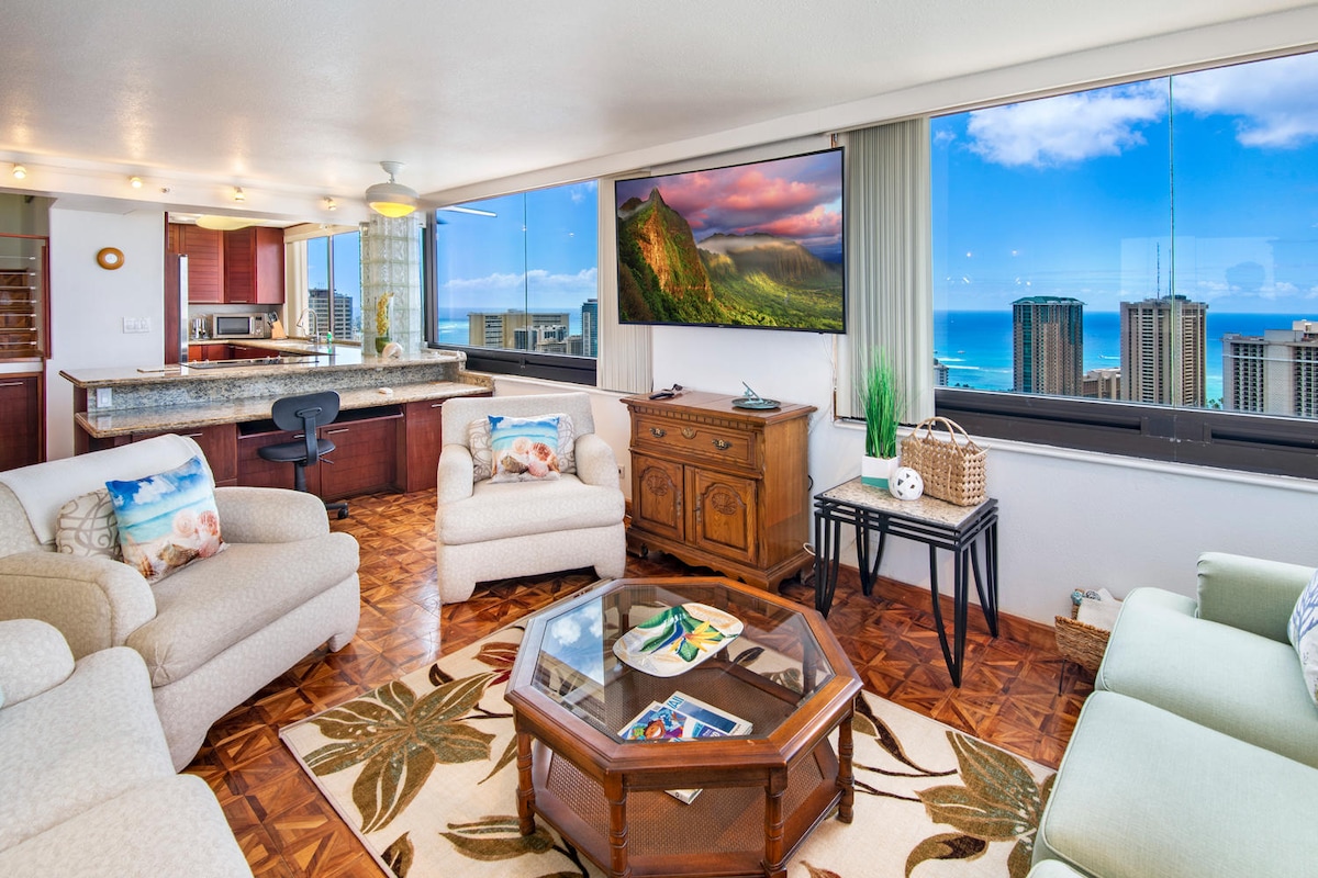 A bright living area is showcased with large windows offering expansive ocean and city views. Comfortable seating is arranged around a wooden coffee table on a floral rug. A mounted TV and a dining area can be seen in the background, complementing the warm wood accents.