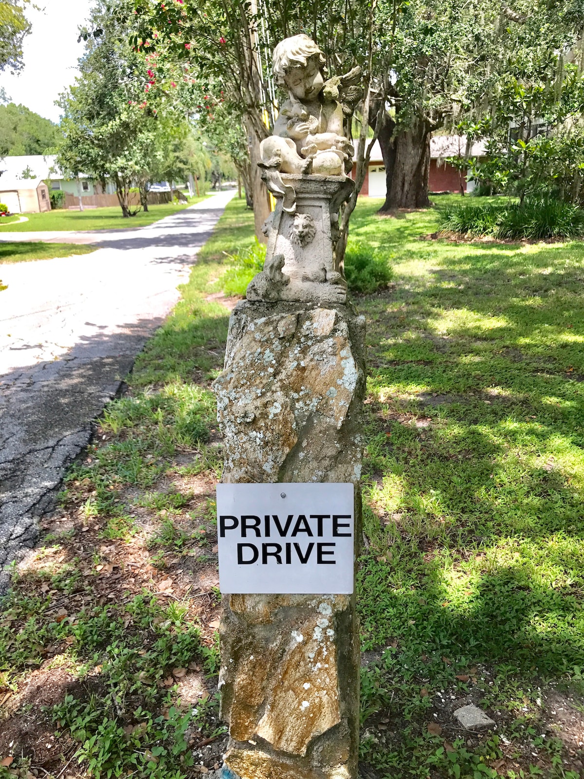 A stone marker stands at the entrance of a private drive, featuring a carved statue of a child. A clear sign displaying 'PRIVATE DRIVE' is affixed to the stone, surrounded by greenery and a glimpse of the residential road ahead.