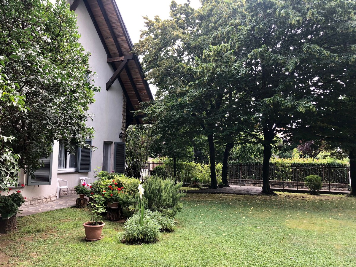 A spacious garden area surrounds a villa, characterized by lush greenery and mature trees. Flower pots are placed throughout, lending a touch of color to the outdoor space. The neatly maintained lawn offers a serene setting, with a fence visible in the background.