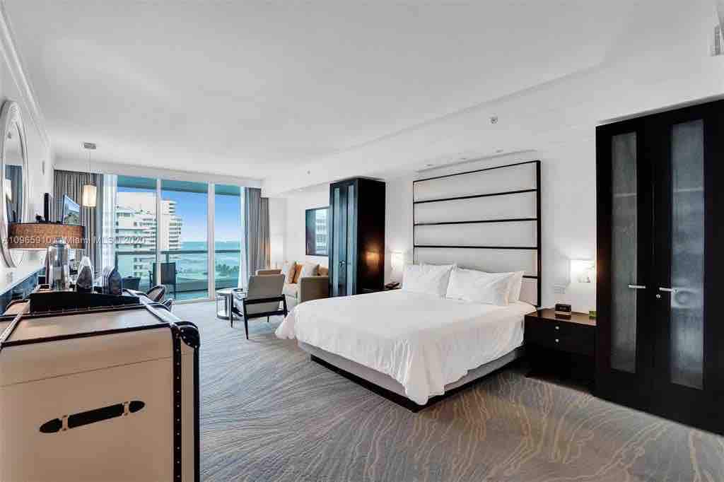 The spacious room features a king size bed with fresh linens, a seating area with armchairs, and a balcony with ocean views. Large windows allow natural light while offering a glimpse of the beach. Storage and a small kitchenette are available along the wall.