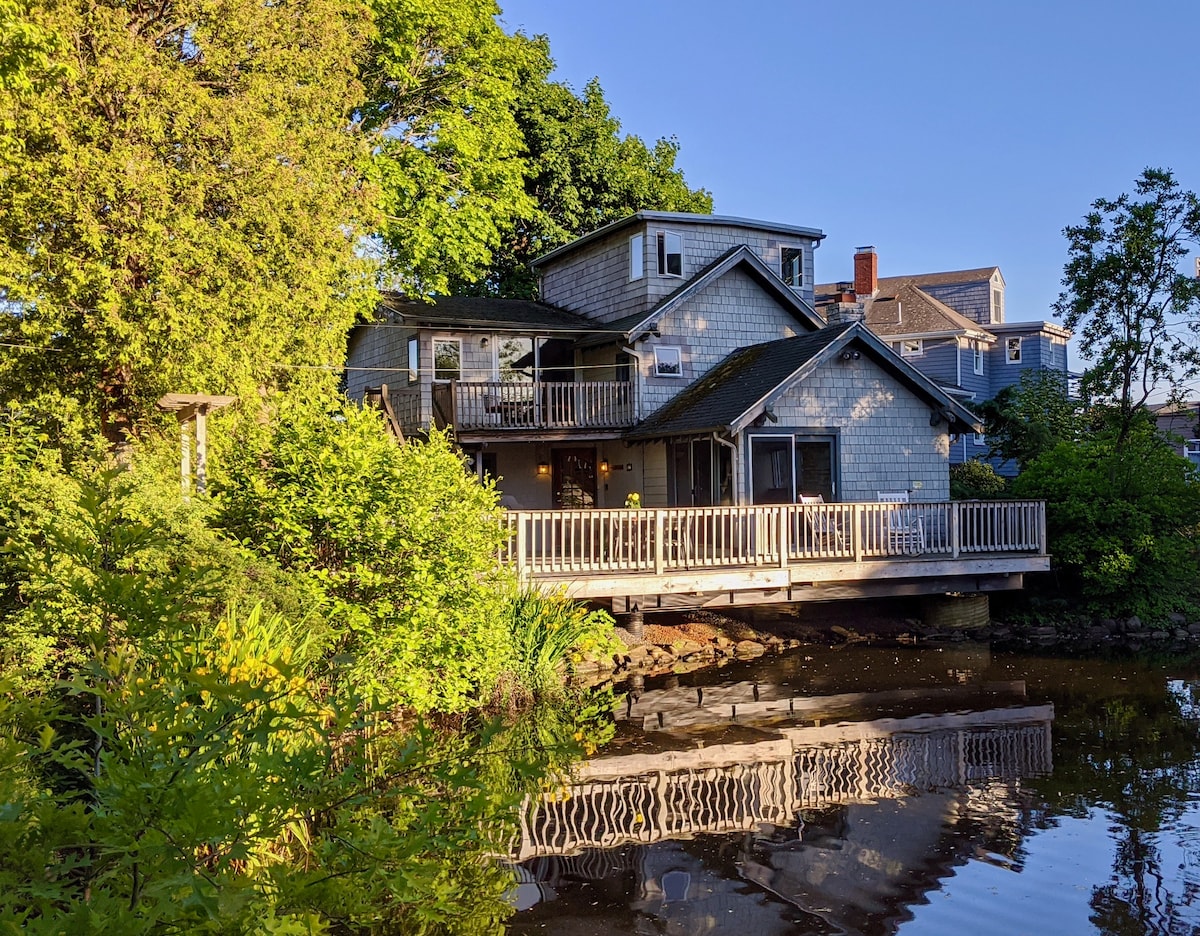 The house is set beside a tranquil pond, featuring a spacious deck surrounded by greenery. The reflection of the structure on the water adds to the serene atmosphere. The shingle-style exterior complements the natural surroundings, creating a harmonious blend with the landscape.