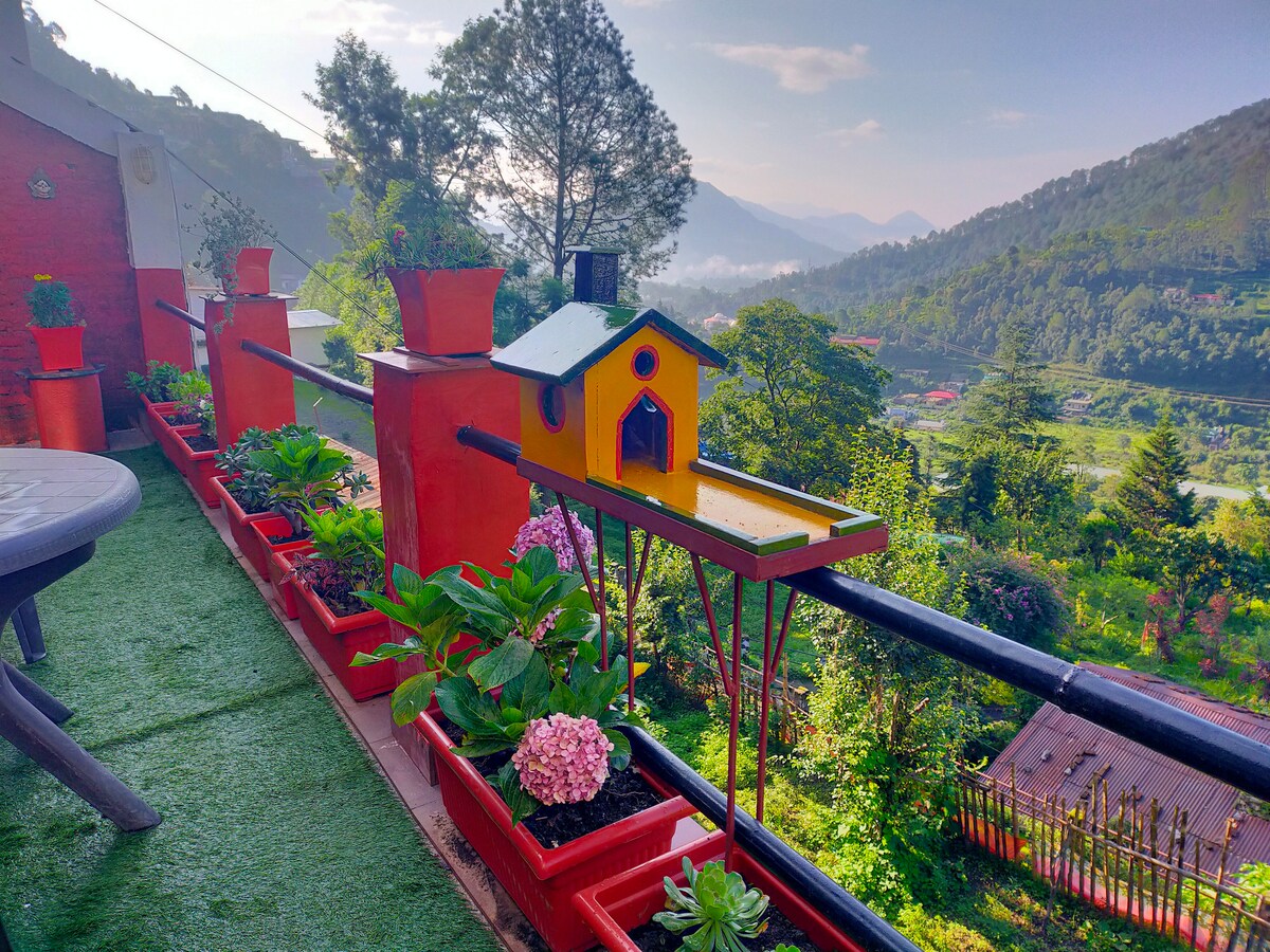 A vibrant balcony garden is adorned with colorful planters filled with various plants, including flowers and succulents. A quaint birdhouse is positioned amongst the greenery, with scenic views of the mountains and valley in the background, creating a serene and inviting outdoor space.