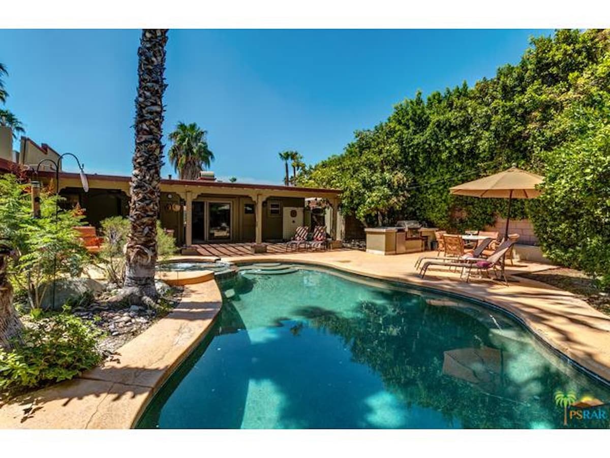 A serene outdoor area features a saltwater pool surrounded by lush greenery and palm trees. Comfortable lounge chairs are positioned beside the pool, while an umbrella provides shade over a dining space. The outdoor kitchen and patio area are visible in the background.