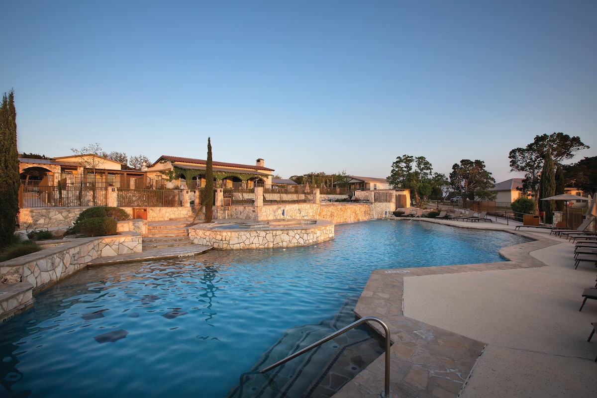 A spacious outdoor pool area is highlighted, surrounded by natural stone walls and landscaping. Lounge chairs are placed along the edge, providing seating options. The tranquil water reflects the evening sky, offering a calming atmosphere.