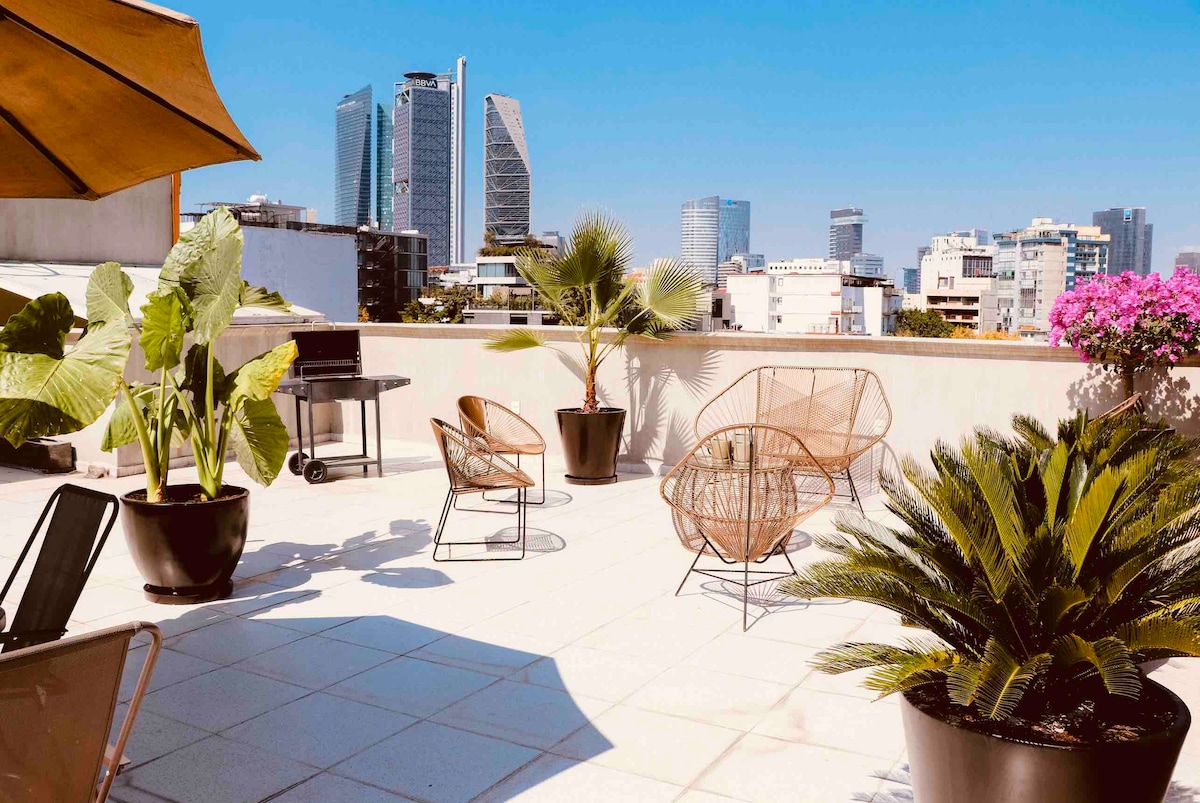 A spacious rooftop area features modern seating arrangements, surrounded by decorative potted plants. A grill is available for use, and vibrant flowers add color against the backdrop of city skyscrapers and a clear blue sky.
