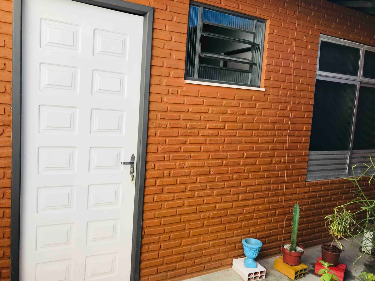 An exterior view of the entrance features a solid white door against a textured orange brick wall. A small window is visible beside the door, with metal grilles. Potted plants, including a cactus, add greenery to the space.