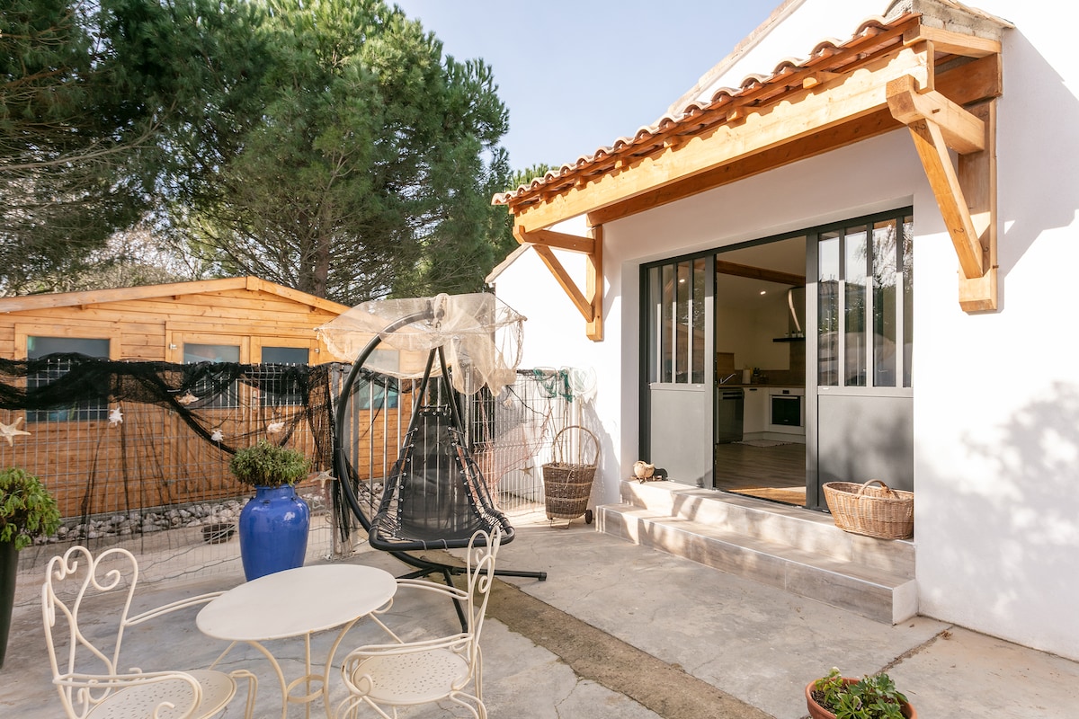 An outdoor space is visible, featuring a swinging chair and a small round table with two chairs. The entrance showcases large glass doors leading into a bright interior. A wooden structure can be seen in the background, surrounded by greenery.