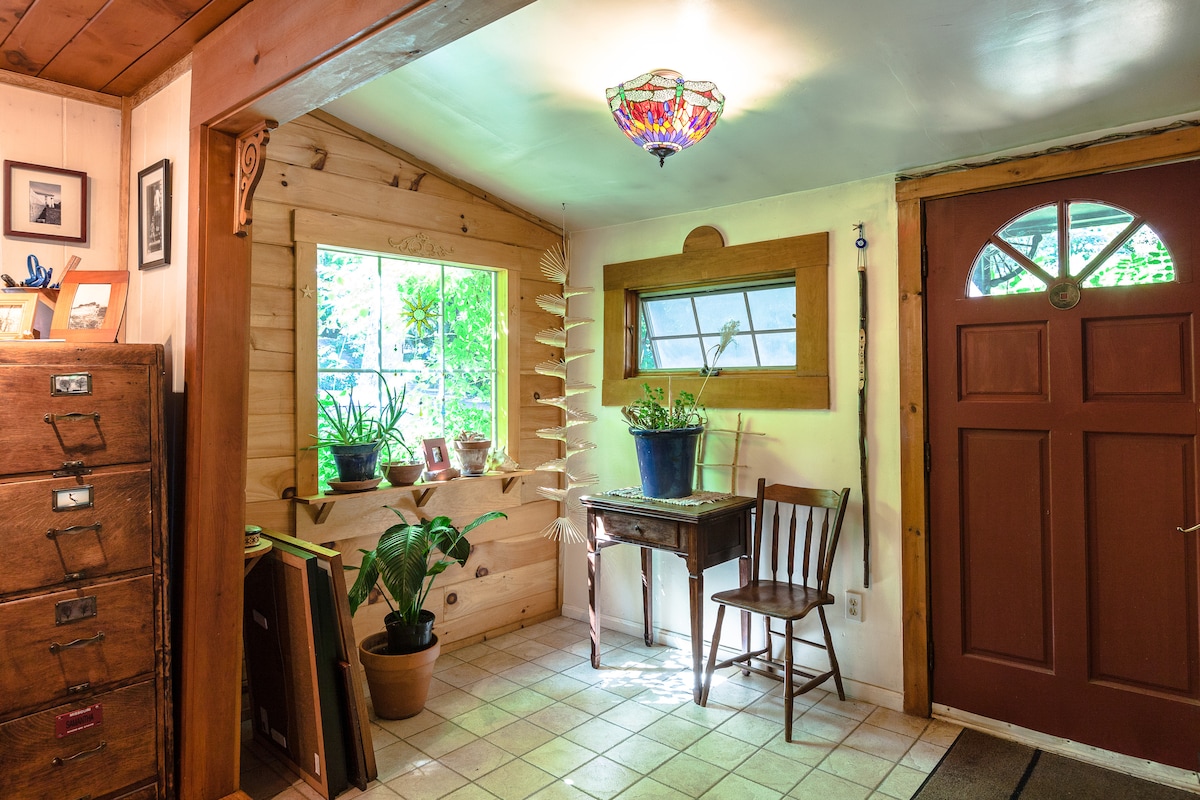The entrance showcases a wooden door with a glass insert, framed by a rustic interior. Sunlight filters through a large window adorned with plants, enhancing the natural ambiance. A small table and chair sit beside the window, complemented by an artistic light fixture overhead.