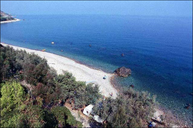 A serene coastline is depicted, showcasing a pebbled beach that gently meets the calm blue waters. Lush greenery is present along the shoreline, while a few boats can be seen on the water. The distant landscape includes rocky formations and hints of coastal structures.