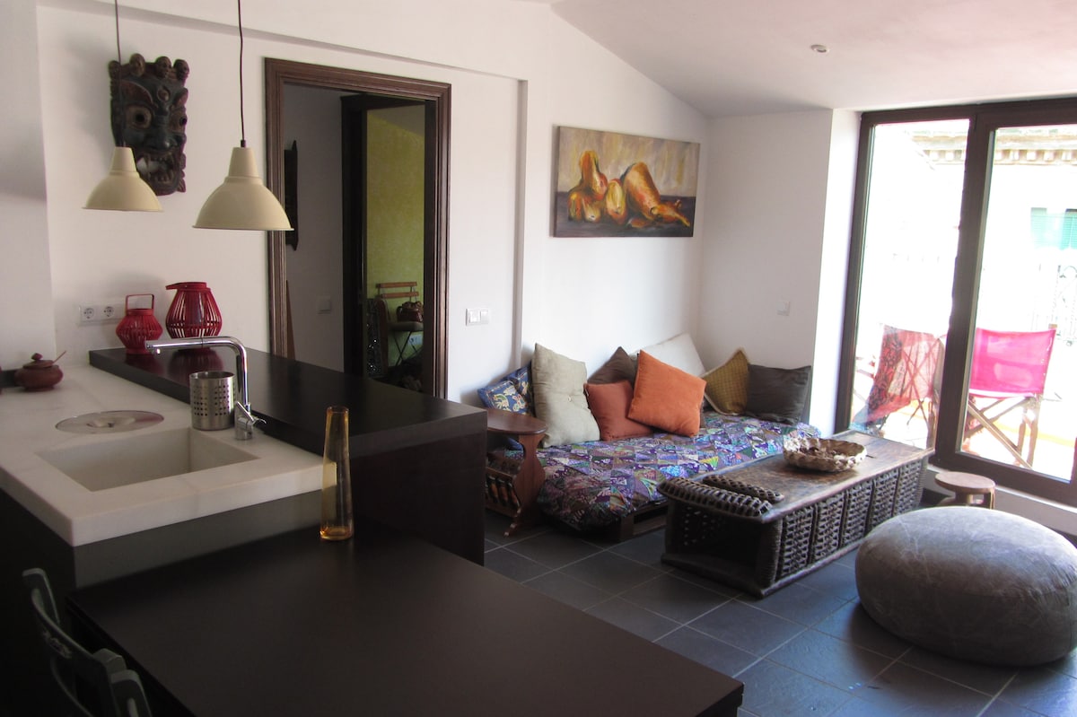 An open living area is presented with a comfortable seating arrangement featuring colorful cushions on a patterned mat. A low wooden table is placed in the center, accompanied by modern pendant lights above. Large glass doors provide natural light and connect to an outdoor space.