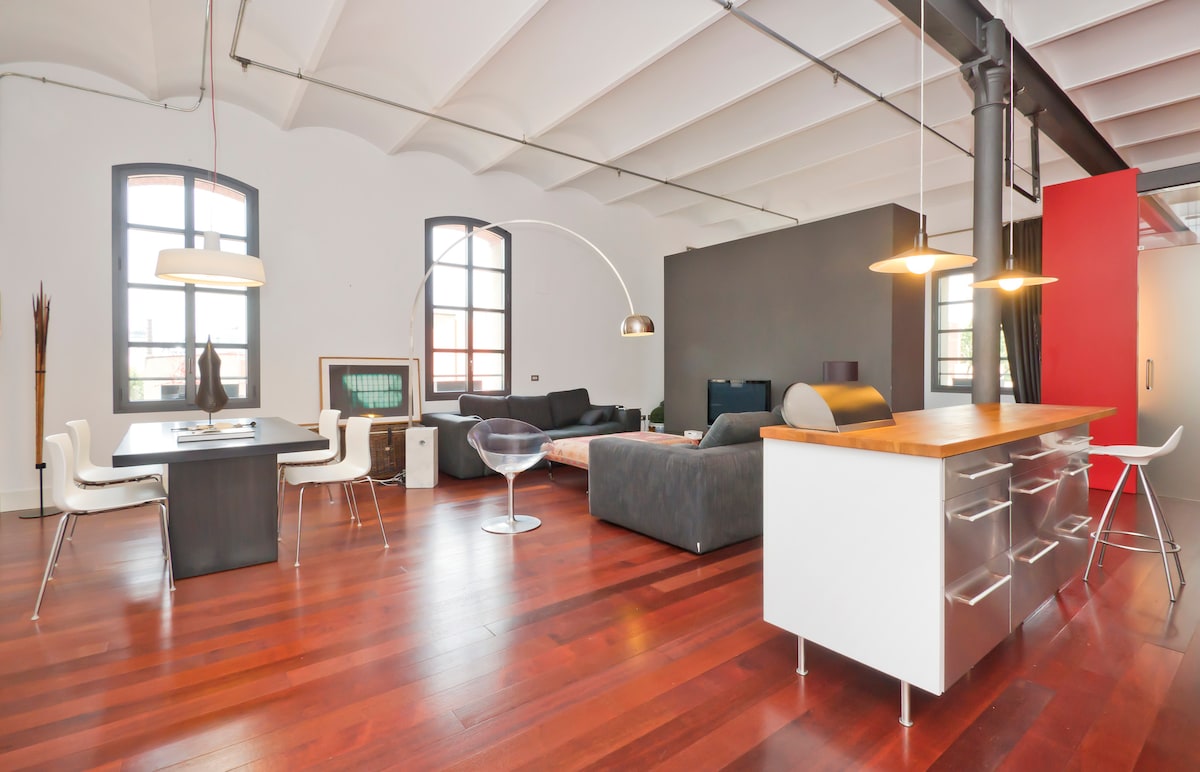The spacious living area highlights a modern design with large windows allowing abundant natural light. A dining table with white chairs sits near a sleek kitchen island, while a comfortable sofa is positioned to face a lively seating area. Warm wooden flooring complements the decor.