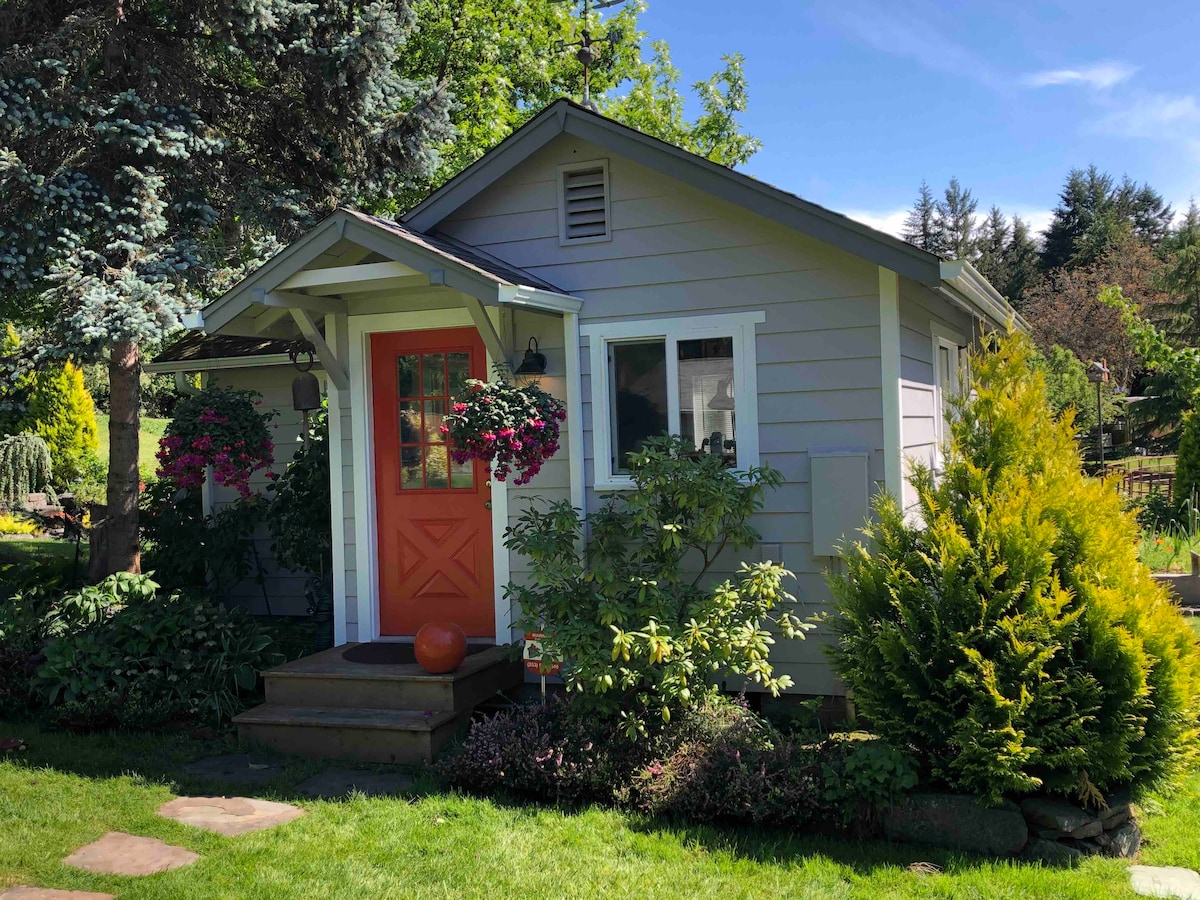 A charming guesthouse is framed by lush greenery and colorful flowering plants. It features a welcoming orange front door and steps leading up to the entrance. The well-maintained lawn adds to the serene setting, complemented by surrounding trees.