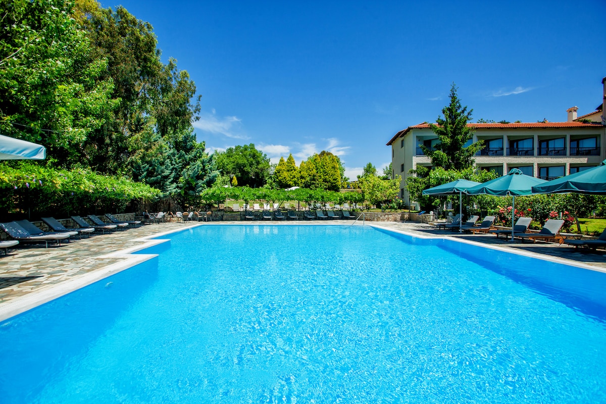 A large swimming pool is surrounded by lush greenery, with sun loungers positioned around its perimeter. Umbrellas offer shade, and the vibrant blue water reflects the clear sky. A building with multiple stories is visible in the background.