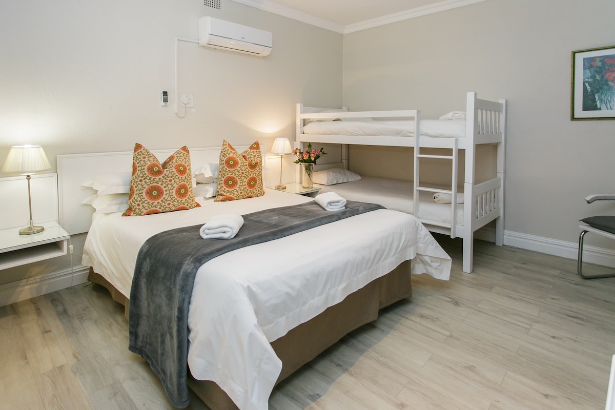 A room equipped with a spacious queen bed adorned with decorative pillows and a cozy throw blanket. A bunk bed is positioned nearby, suitable for both an adult and a child. Soft lighting is provided by bedside lamps, and the air conditioning unit is mounted on the wall.