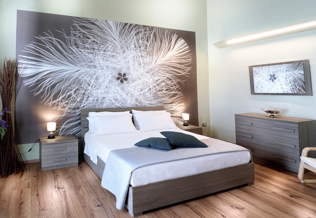 A spacious bedroom features a large double bed with crisp white bedding and two side tables with lamps. A striking wall art piece in monochrome enhances the room's aesthetic. Warm wooden flooring adds to the inviting atmosphere, while natural light fills the space.