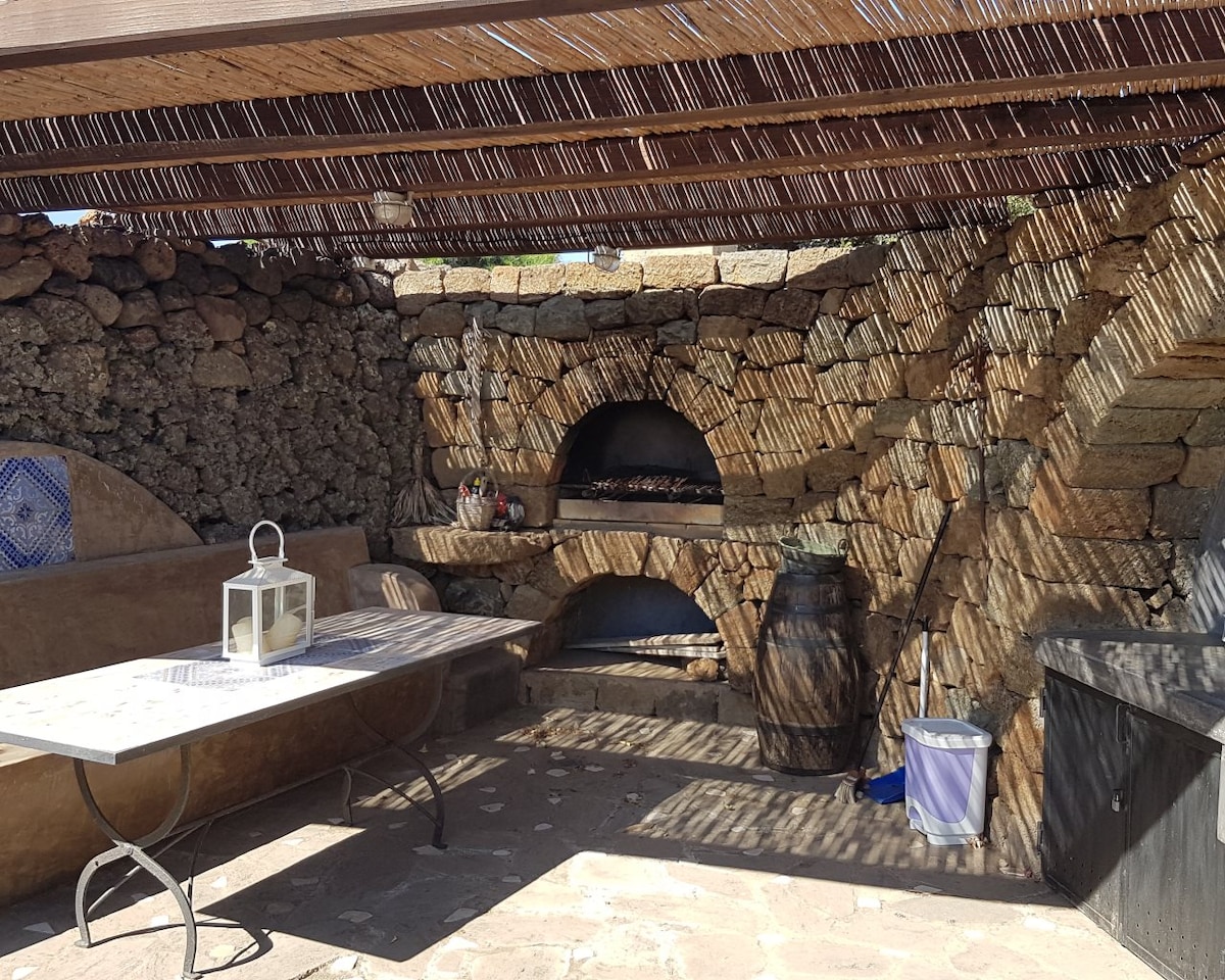 An outdoor space features a stone barbecue area with a built-in grill, surrounded by rustic stone walls. A long table and bench provide seating, while a lantern and a barrel add to the rustic charm. Natural light filters through a reed roof, casting shadows on the ground.
