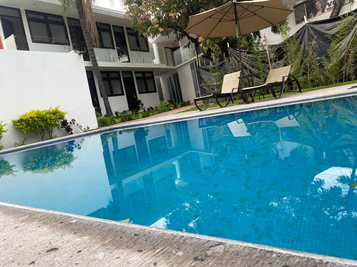A tranquil swimming pool reflects the surrounding greenery and buildings. Two lounge chairs sit at the poolside, shaded by a large umbrella. The setting is framed by a well-manicured lawn and modern architecture in the background.