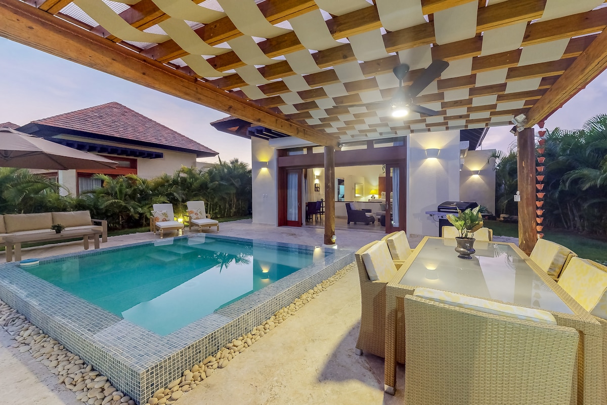 The private outdoor area features a plunge pool surrounded by a stone border and a shaded patio with a dining table. Comfortable seating options are arranged nearby, and lush greenery is visible in the background, creating a serene and relaxing environment.