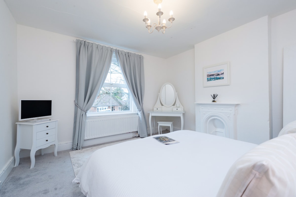 A bright bedroom features a spacious bed dressed in crisp white linens. A large window is dressed with light grey curtains, allowing natural light to fill the room. A small television and a dresser are positioned against the wall, complementing the elegant decor.