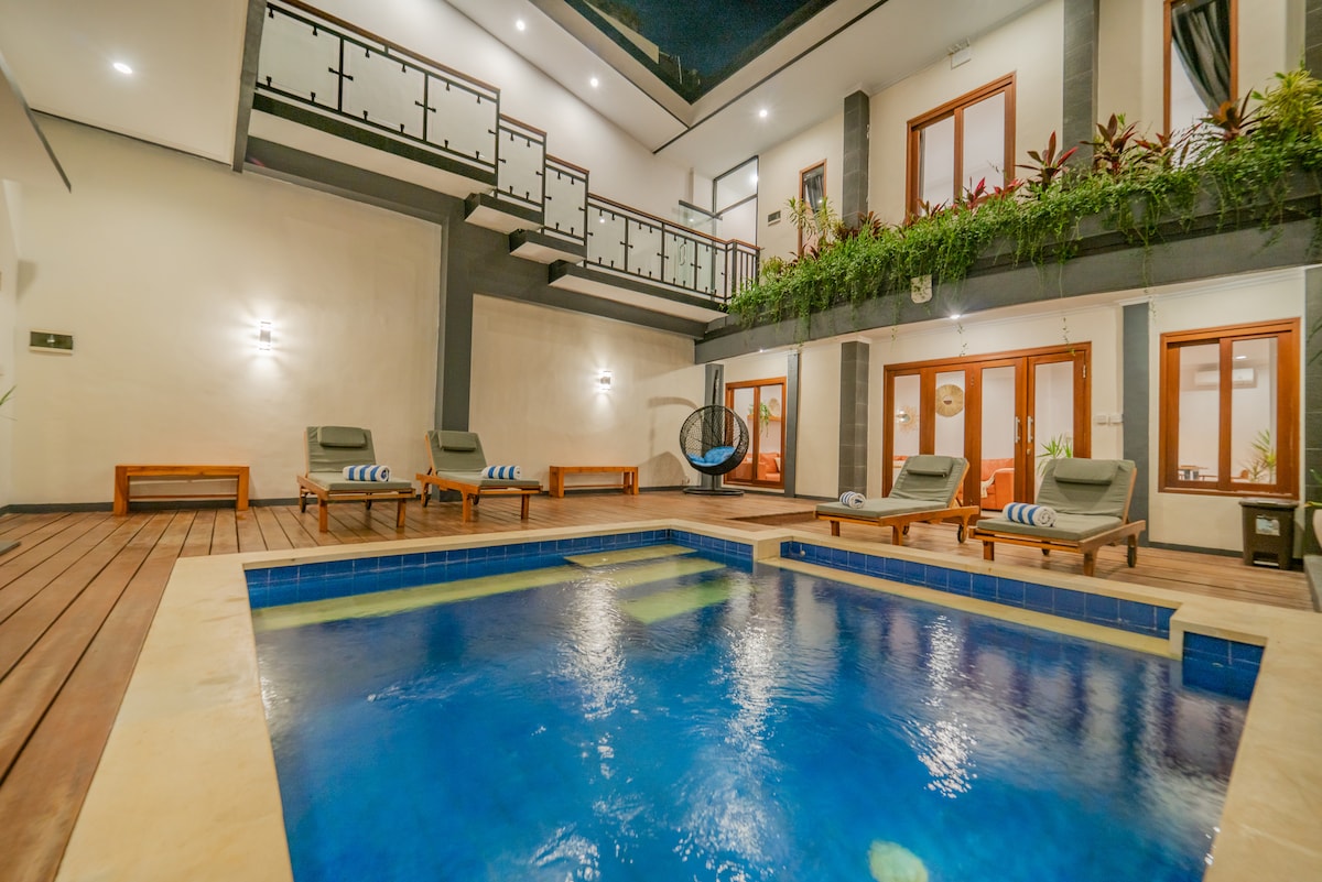 The private pool area features a rectangular pool surrounded by wooden decking. Lounge chairs with towels are positioned nearby. A wall of greenery and large windows enhances the space, while the second floor balcony provides additional seating options.