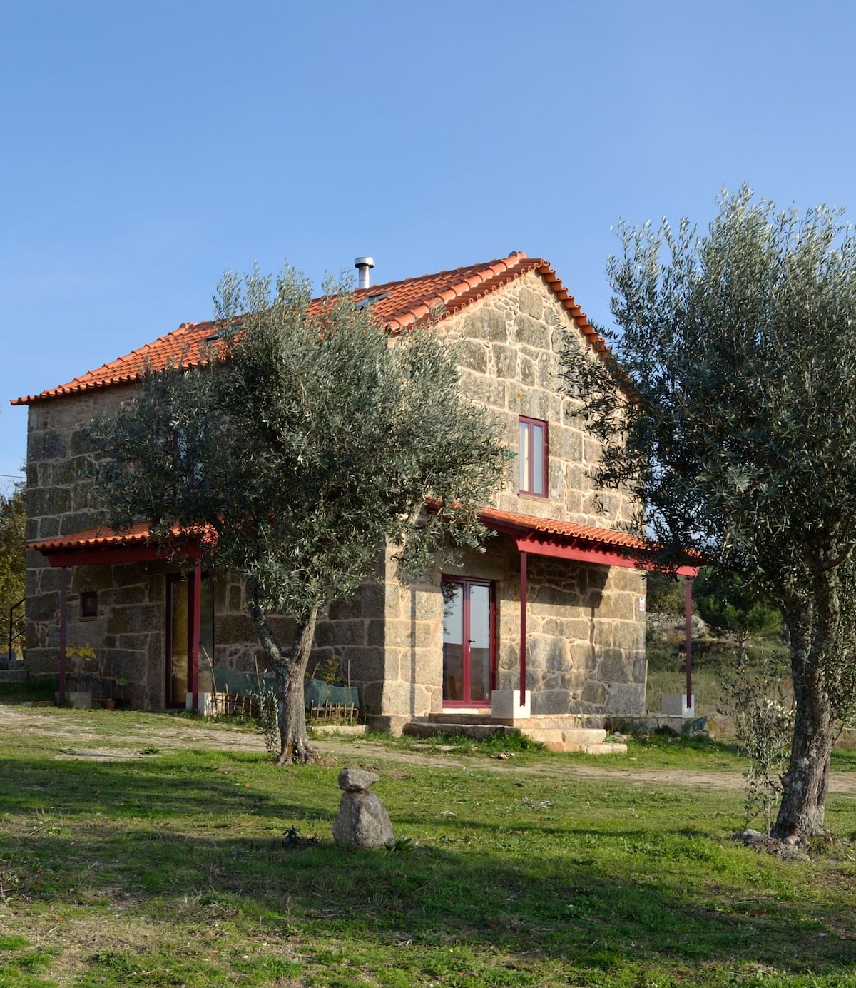 A rustic stone cottage is framed by two olive trees, set against a clear blue sky. The cottage features a red-tiled roof and large windows, allowing natural light to fill the interior. A well-manicured lawn surrounds the structure, enhancing the rural ambiance.