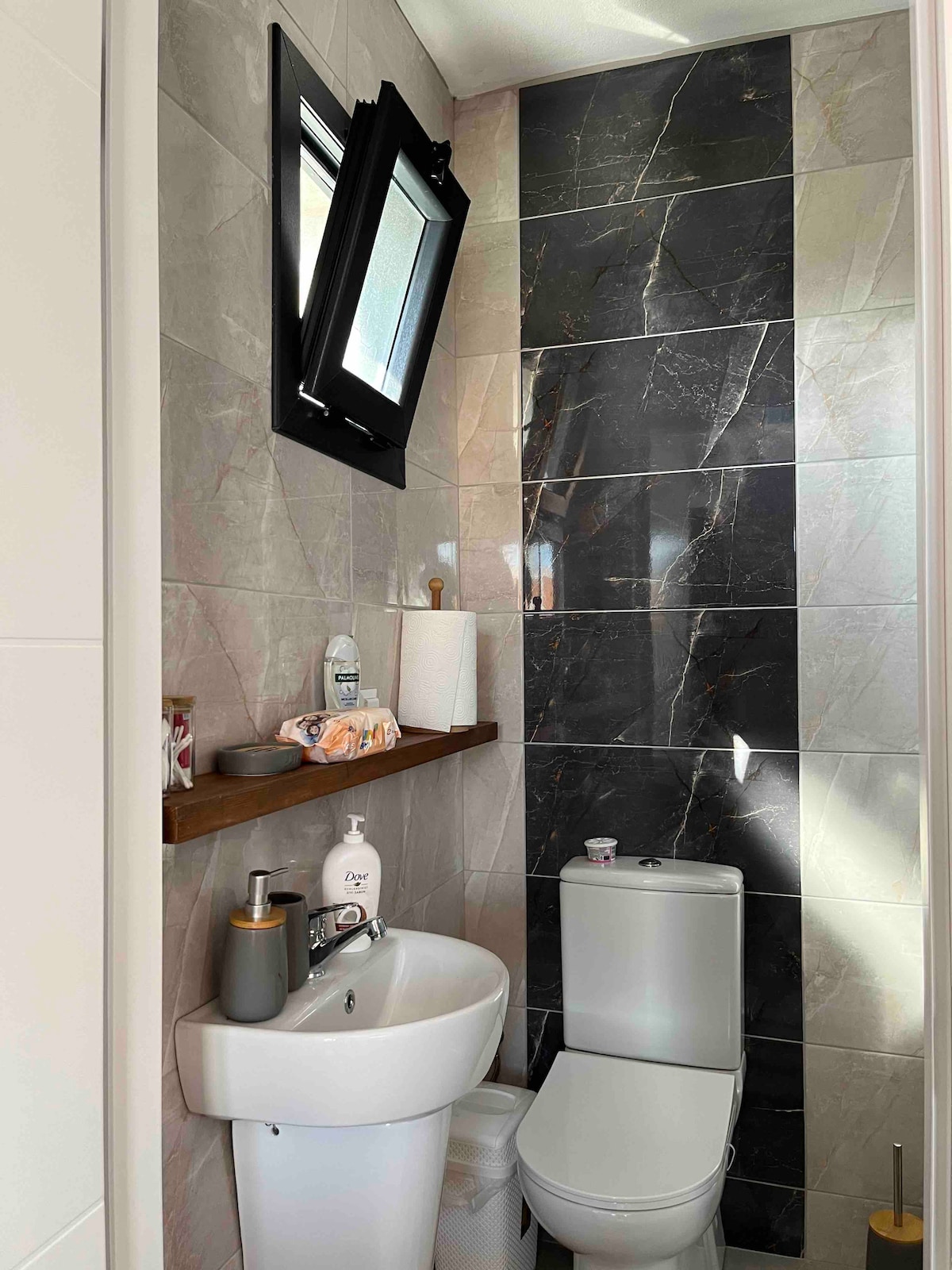 A clean, modern bathroom features elegant dark and light marble tiles, complemented by a wooden shelf holding toiletries. A compact white toilet and sink are positioned for convenience. A small window allows natural light to enhance the space.
