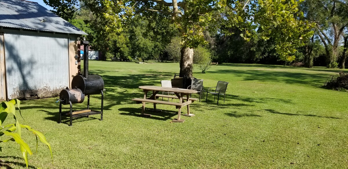 A well-maintained outdoor area features a picnic table surrounded by four chairs. A grill stands nearby under a large shade tree, with a manicured lawn stretching expansively in the background. The structure's exterior shows a rustic appearance.