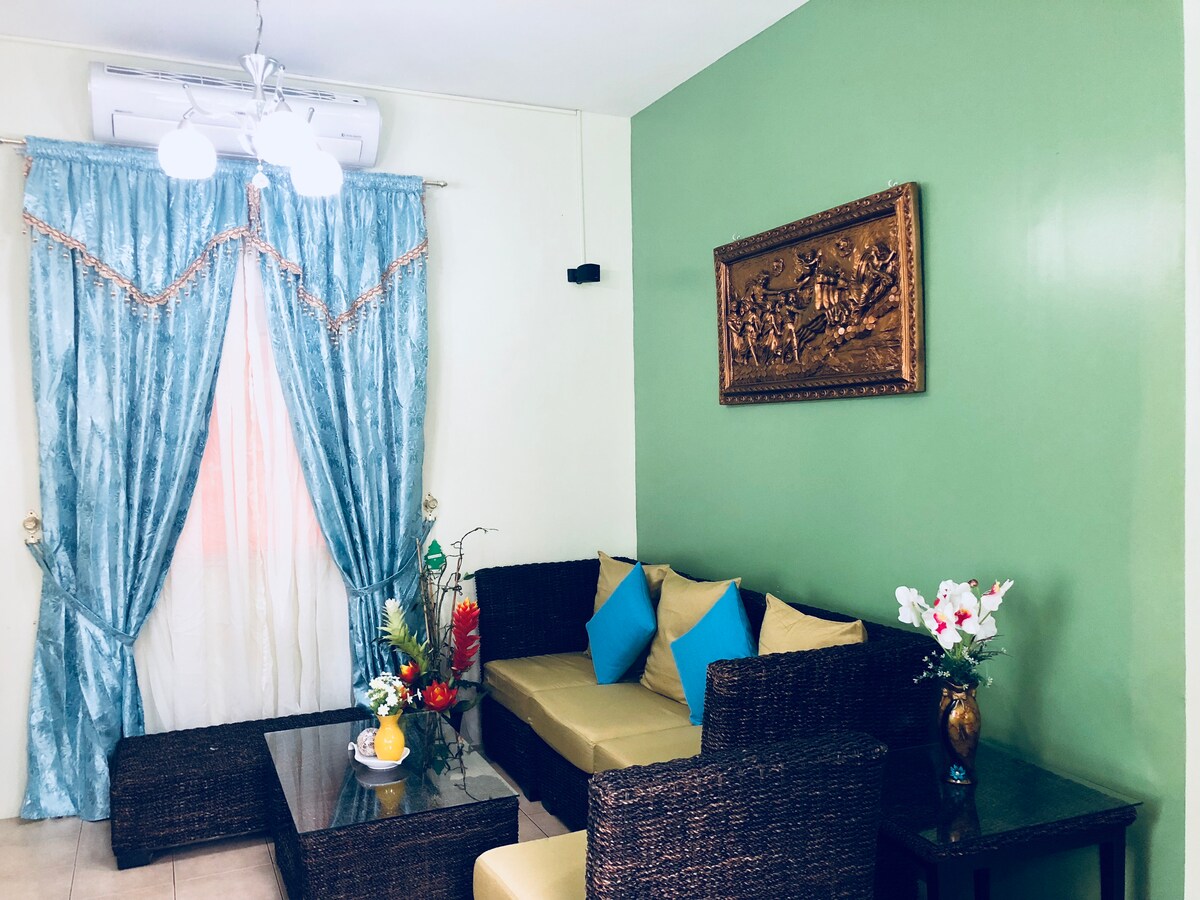 A cozy living room features a dark wicker sofa adorned with yellow cushions, situated around a glass coffee table. Light blue curtains frame a window, while a wall art piece adds a touch of character. Decorative plants and flowers enhance the inviting atmosphere.