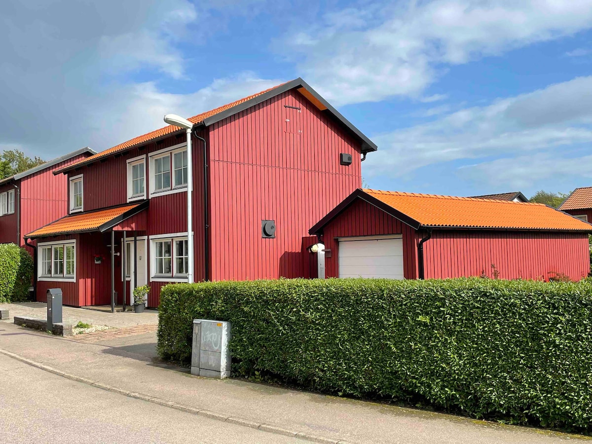 A two-story red wooden house with a distinctive orange-tiled roof is situated in a well-kept neighborhood. Large windows provide ample natural light, and a neatly trimmed green hedge lines the front, creating a welcoming entrance. The garage is positioned to the right of the home.
