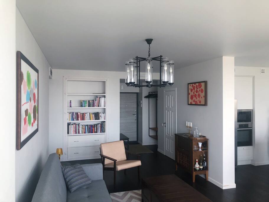 The living room as seen from the window side and featuring the sofa, one of the chairs, the bar, the bookshelves and the entrance to the apartment.