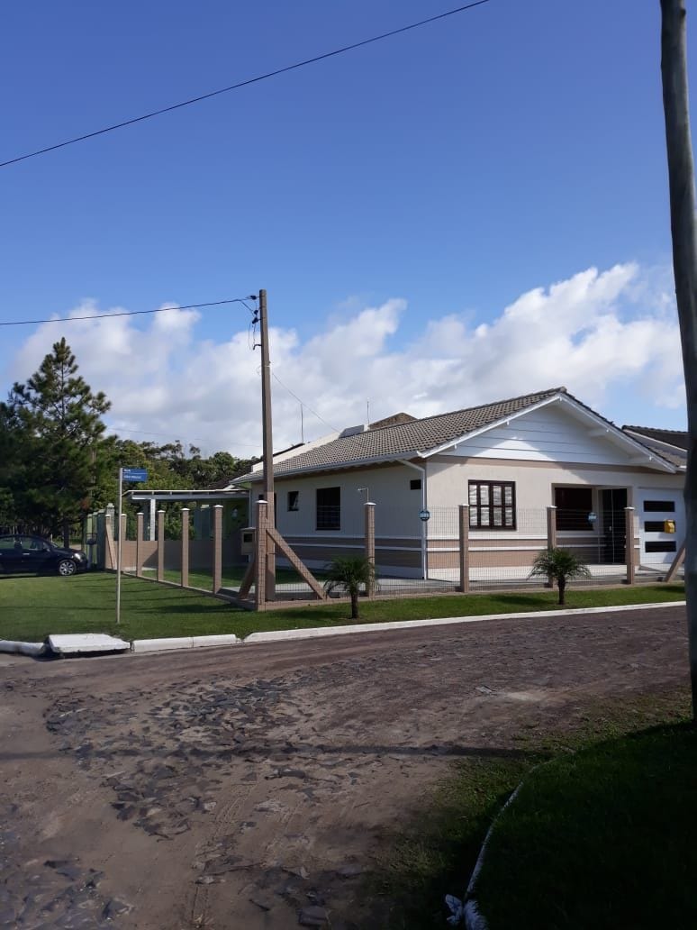 A single-story house is seen from the street, featuring a covered porch and a neatly landscaped yard. The exterior displays a combination of light and dark colors, with large windows and a sloped roof. A driveway is visible, as are surrounding trees and a clear blue sky.