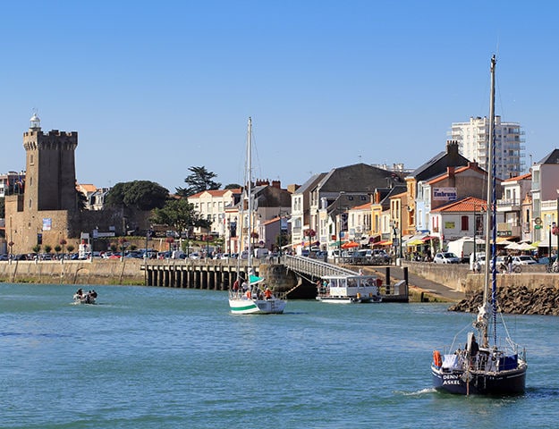 A view of a serene harbor is presented, featuring several boats gently bobbing in the water. A historic tower stands prominently on the left, surrounded by charming buildings that line the waterfront. The clear blue sky reflects on the tranquil surface of the harbor.