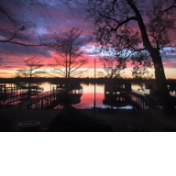 A serene lakefront view captures a vibrant sunset, showcasing hues of pink and purple reflected on calm waters. Dock structures are visible along the shoreline, and silhouettes of trees frame the scene, providing a natural backdrop.