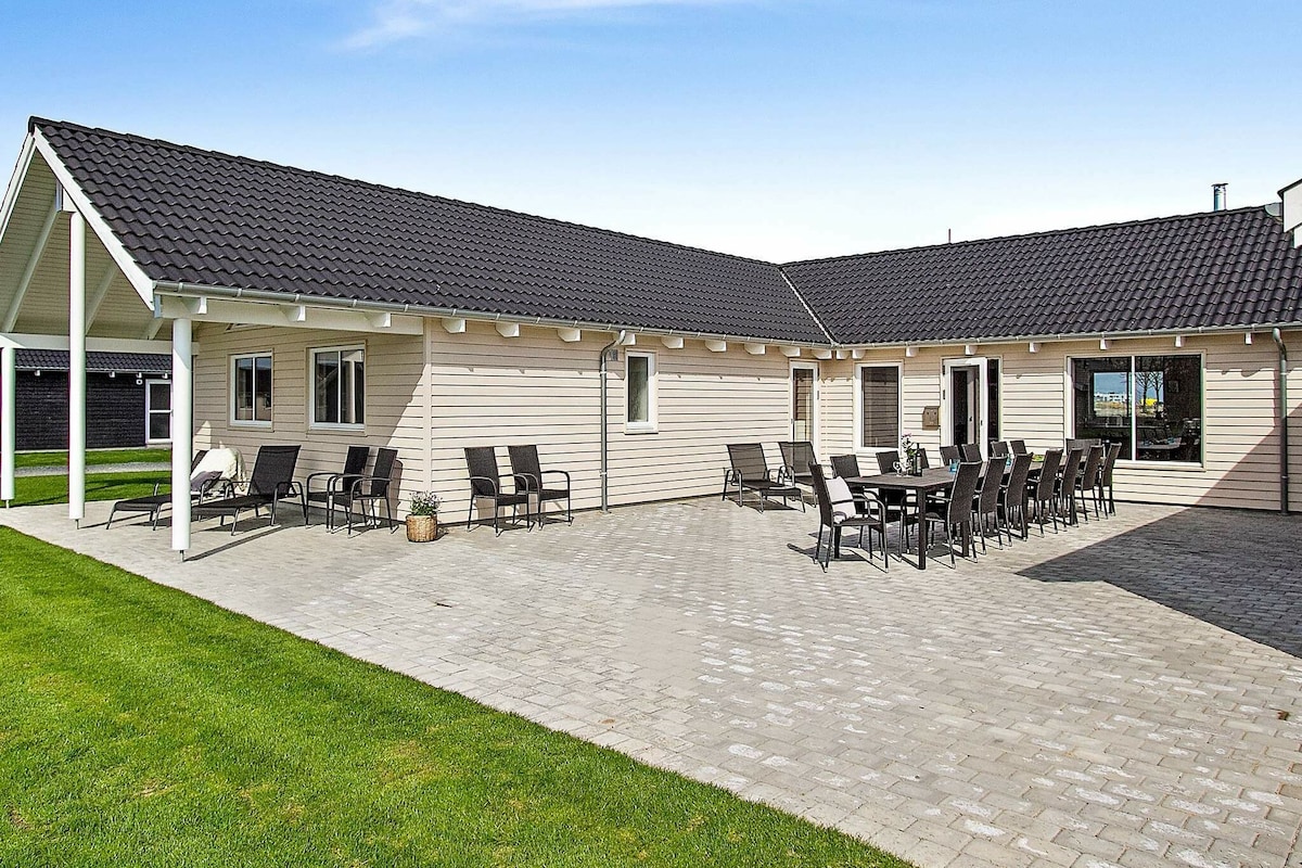 A spacious outdoor patio is featured, paved with square stones and surrounded by a lush green lawn. A large dining table is set with multiple chairs, along with several lounge chairs positioned for relaxation. The structure is designed with a sloping roof, enhancing the area’s ventilation.
