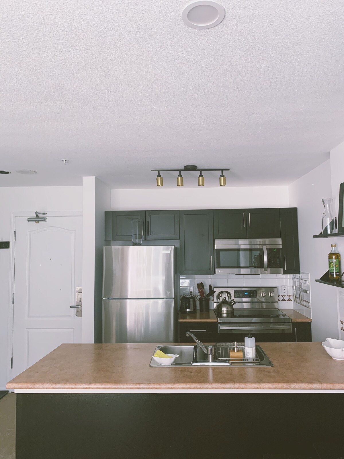 A modern kitchen features sleek black cabinetry and stainless steel appliances, including a refrigerator, oven, and microwave. A light fixture with multiple bulbs hangs from the ceiling, and a kitchen island with a sink provides additional workspace. Open shelving displays kitchen essentials.