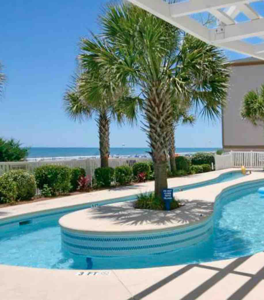 An outdoor pool is surrounded by tall palm trees and lush greenery. The pool features a lazy river design, with gentle curves inviting relaxation. The Atlantic Ocean is visible in the background, under a clear blue sky, providing a serene coastal environment.