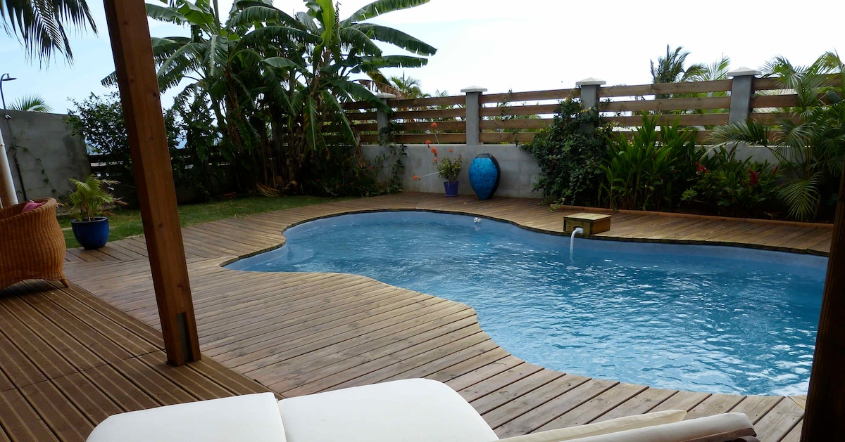 A serene outdoor space features a wooden deck surrounding a curved pool. Lush green plants and palm trees provide a tropical ambiance, while a blue decorative pot adds a pop of color. A comfortable lounge chair is positioned for relaxation near the water.