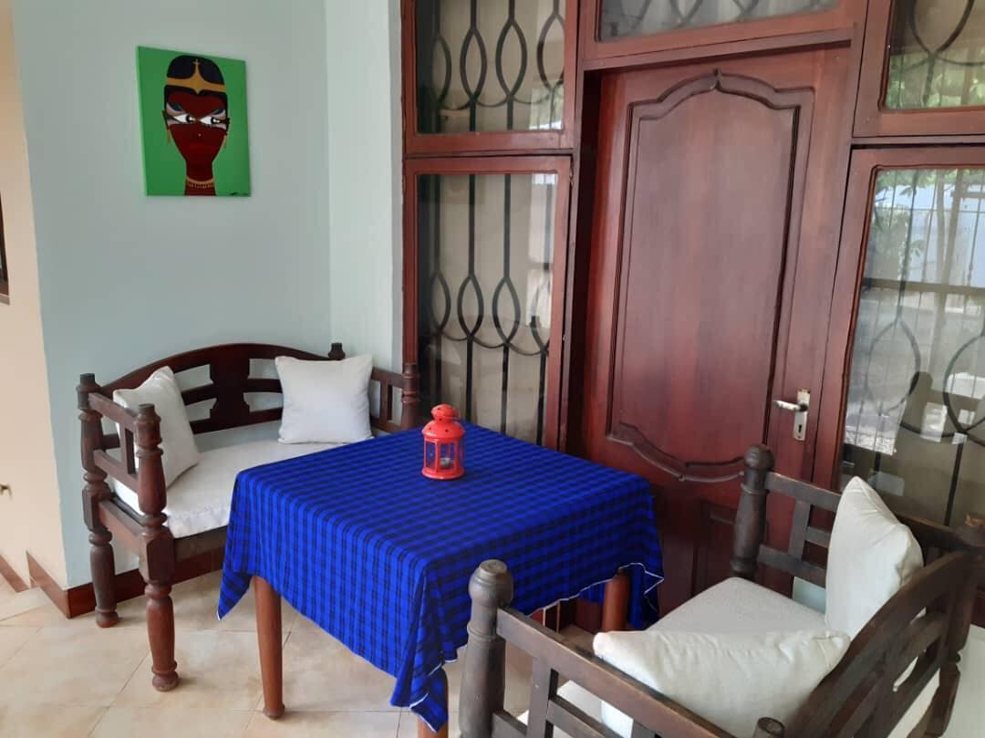 A welcoming seating area is featured, with two wooden benches adorned with white cushions facing a small table draped in a blue checkered cloth. A red lantern sits at the center of the table, and large glass doors provide ample natural light.