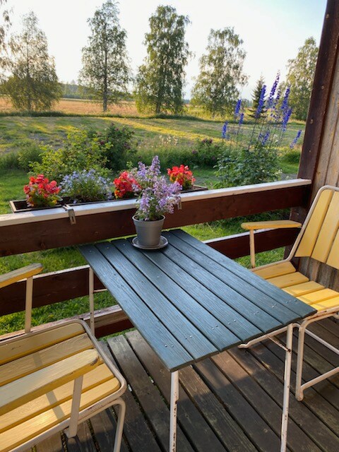 A small covered patio features a table and two striped chairs. A potted plant sits on the table, surrounded by colorful flowers in window boxes. A picturesque landscape with trees and fields is visible in the background.