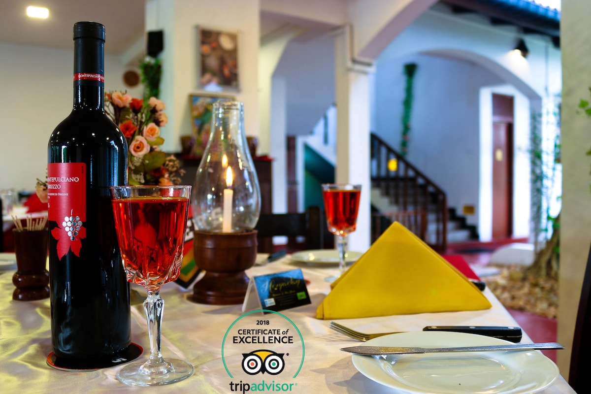A dining table is elegantly set with two glasses filled with a vibrant red beverage, accompanied by a bottle of wine. A yellow napkin adds a bright touch next to decorative flowers. The background reveals subtle architectural details and a warmly lit staircase.