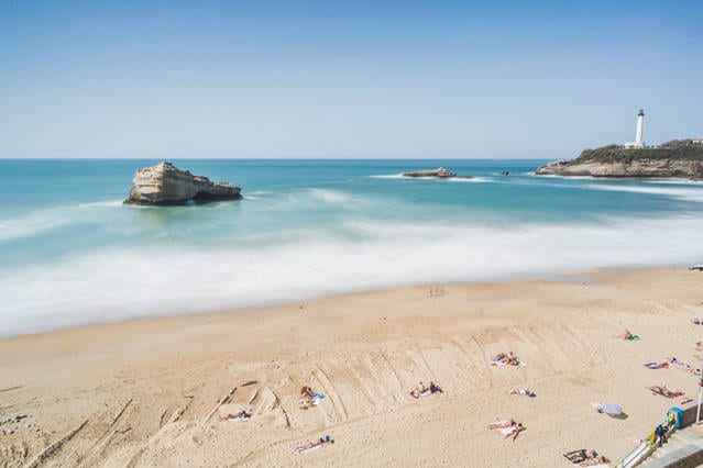 A calm beach scene features soft, golden sand extending toward gentle waves. In the distance, a lighthouse stands on a rocky outcrop, while the horizon showcases a clear blue sky, enhancing the relaxing coastal atmosphere.