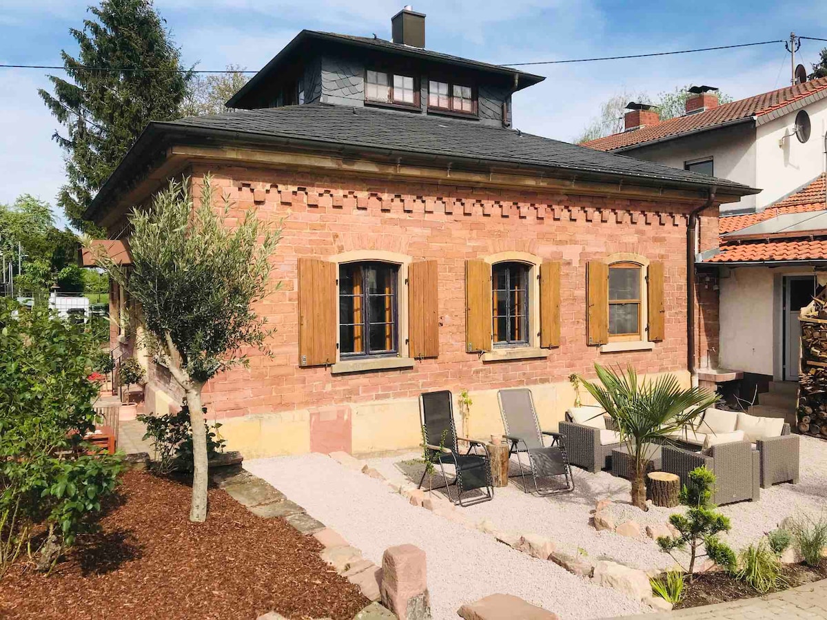 The exterior of a charming historic house is depicted, featuring exposed brick walls and brown wooden shutters. Two lounge chairs are positioned on a gravel area surrounded by small plants and rocks, with a palm tree adding greenery. A tree is visible in the background.