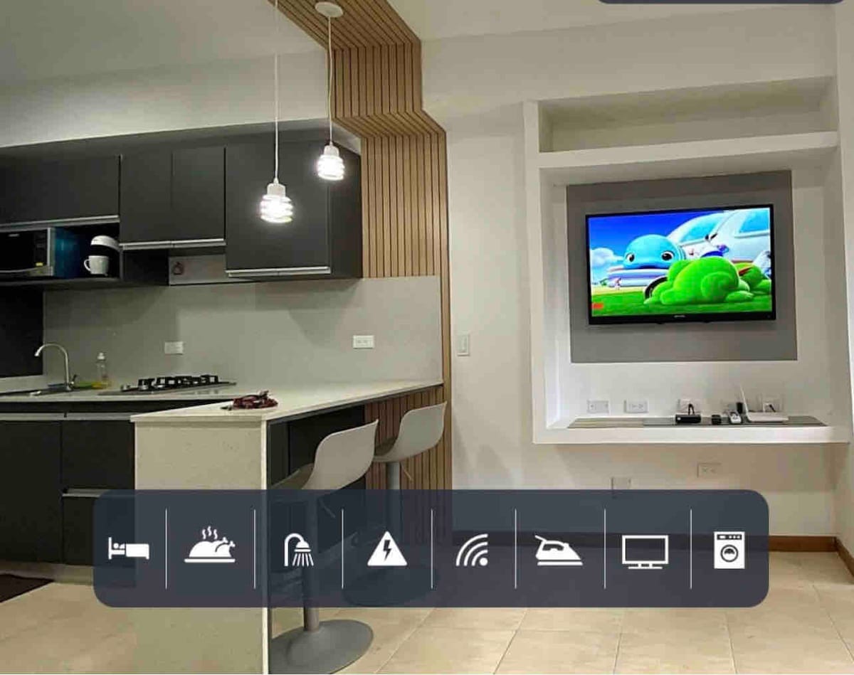 A modern kitchen and living area is showcased, featuring sleek cabinetry and a spacious countertop. Two bar stools are positioned at the kitchen island. A wall-mounted television displays a colorful cartoon. Soft lighting is provided by pendant lights above the island.