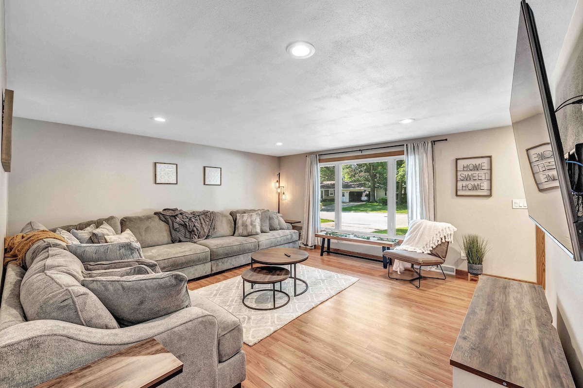 A spacious living room features a large, sectional sofa, inviting relaxation. A round coffee table sits in front, and soft lighting enhances the atmosphere. Natural light filters through the window, which frames a view of the front yard. A comfortable chair adds additional seating.