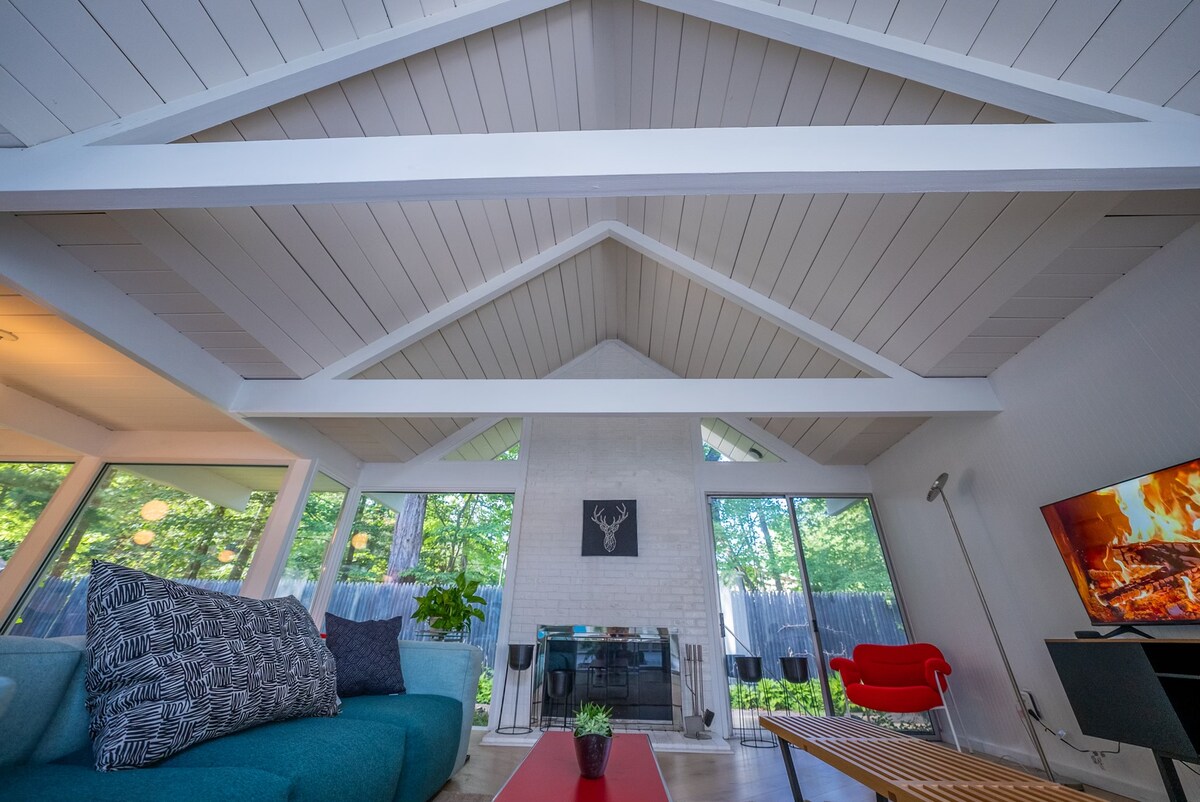 A spacious living area features vaulted ceilings, showcasing a wooden beam structure. Large windows allow natural light to fill the space, highlighting a modern fireplace and minimalist furnishings, including a teal sofa and a red accent chair. A small plant sits on the coffee table.