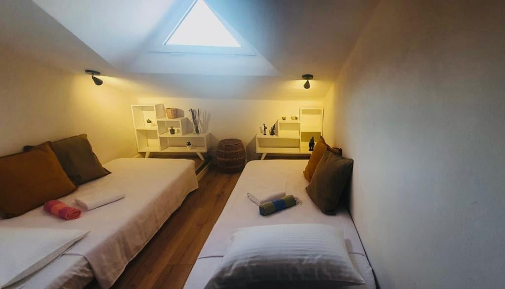 A bright sleeping area features two spacious beds with neutral linens and assorted pillows. A low table is adorned with decorative items, and natural light is welcomed through a skylight. Soft lighting from wall sconces enhances the overall ambiance.